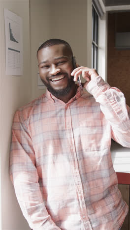 Vertical-video-portrait-of-happy-african-american-casual-businessman-talking-on-smartphone-in-office