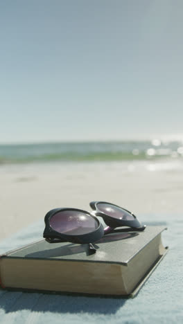 Vertical-video-of-book,-sunglasses-and-towel-on-beach,-in-slow-motion,-with-copy-space