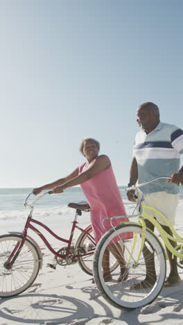 Video-Vertical-De-Una-Pareja-Afroamericana-Caminando-Con-Bicicletas-En-La-Playa,-En-Cámara-Lenta