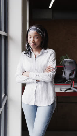 Vertical-video-portrait-of-african-american-casual-businesswoman-by-window,-smiling-in-office