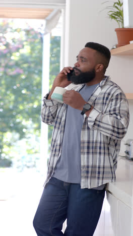 Vertical-video-of-african-american-man-drinking-coffee-talking-on-smartphone-in-kitchen,-slow-motion