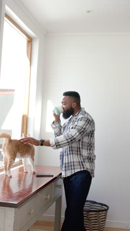 Vertical-video-of-african-american-man-drinking-coffee-and-petting-cat,-slow-motion