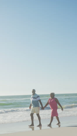 Vertical-video-of-senior-african-american-couple-holding-hands-at-beach,-in-slow-motion