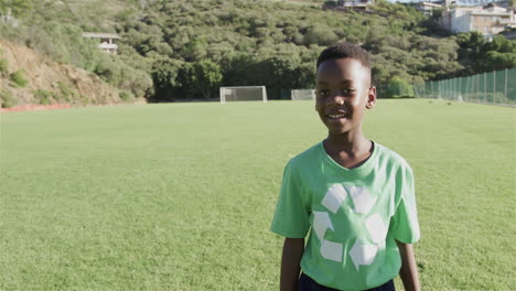 Un-Niño-Afroamericano-Con-Una-Camiseta-Verde-De-Reciclaje-Se-Encuentra-En-Un-Soleado-Campo-De-Fútbol,-Con-Copia-