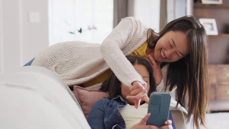 Video-of-happy-asian-mother-and-daughter-sitting-on-sofa-with-smartphone