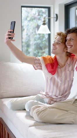 Vertical-video-of-diverse-gay-male-couple-with-lgbt-flag,-using-smartphone-at-home,-slow-motion