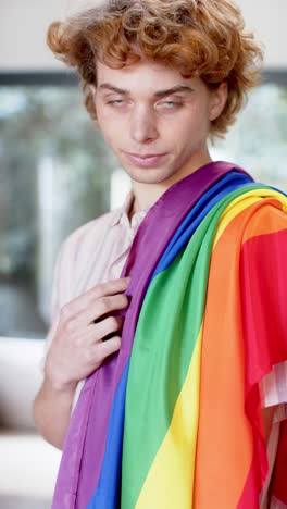 Video-Vertical-Del-Retrato-De-Un-Hombre-Caucásico-Feliz-Sosteniendo-La-Bandera-Lgbt-Del-Arco-Iris-En-Casa,-Cámara-Lenta