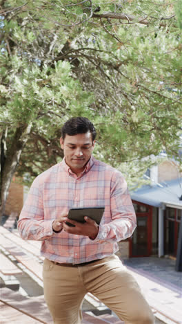 Vertical-video-of-portrait-of-happy-biracial-male-teacher-with-tablet-over-school-in-slow-motion