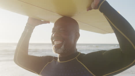 Glücklicher-älterer-Afroamerikanischer-Mann,-Der-In-Zeitlupe-Am-Strand-Spazieren-Geht-Und-Ein-Surfbrett-Hält