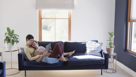 Happy-caucasian-lesbian-couple-lying-on-sofa,-using-tablet-and-laughing-in-sunny-house,-copy-space