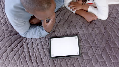 African-american-father-and-son-lying-on-bed-using-tablet-with-copy-space-at-home,-slow-motion