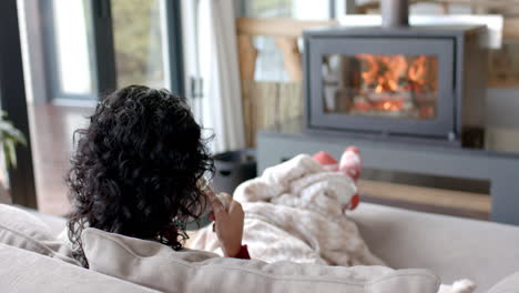 Biracial-woman-drinking-coffee-on-sofa-under-blanket-in-front-of-fireplace-at-home,-slow-motion