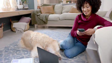 Happy-biracial-woman-with-golden-retriever-dog-using-laptop-and-smartphone-at-home,-slow-motion