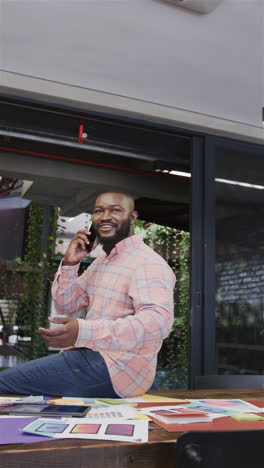 Vertical-video-of-happy-african-american-casual-businessman-sitting-on-office-desk-talking-on-phone