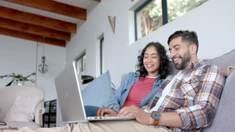 Happy-biracial-couple-sitting-on-sofa-and-using-laptop-at-home,-in-slow-motion