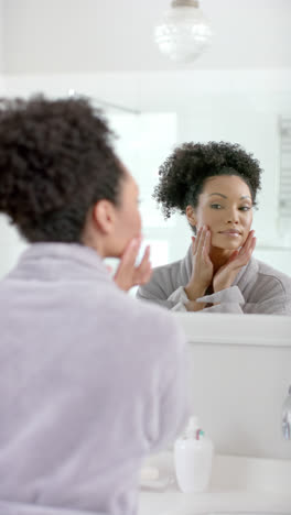 Vertical-video-of-biracial-woman-in-bathrobe-looking-in-mirror-in-bathroom,-slow-motion