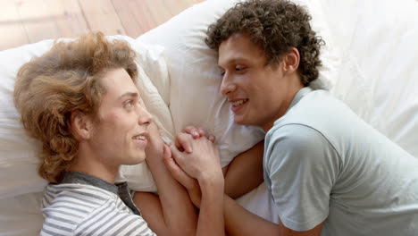 Happy-diverse-gay-male-couple-lying-on-bed-holding-hands-at-home,-slow-motion