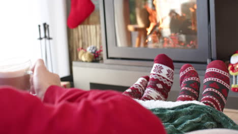 Diverse-gay-male-couple-drinking-cocoa-sitting-on-sofa-by-fireplace-at-home,-slow-motion