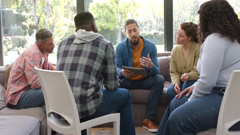 Grupo-Diverso-De-Amigos-Y-Terapeuta-Masculino-Con-Tableta-En-Sesión-De-Terapia-Grupal