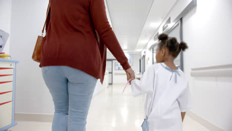 Madre-E-Hija-Afroamericanas-En-Bata-De-Hospital-Cogidas-De-La-Mano-Caminando-En-El-Pasillo,-Cámara-Lenta