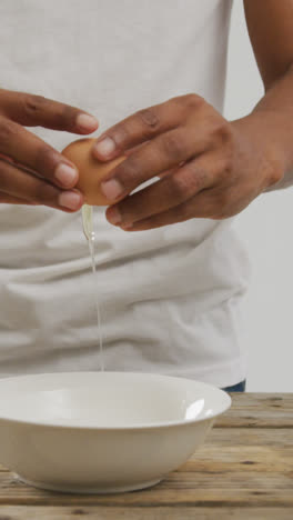 Video-of-biracial-man-cracking-egg-into-white-bowl-on-wooden-surface-and-white-background