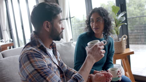 Feliz-Pareja-Birracial-Sentada-En-El-Sofá,-Tomando-Café-Y-Hablando-En-Casa,-En-Cámara-Lenta