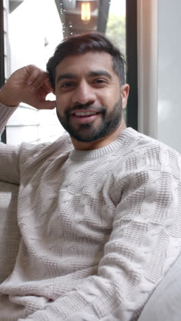 Vertical-video-of-portrait-of-happy-biracial-man-sitting-on-sofa-at-home,-slow-motion