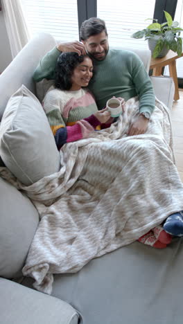 Vertical-video-of-biracial-couple-sitting-on-sofa-embracing-and-drinking-coffee-at-home,-slow-motion