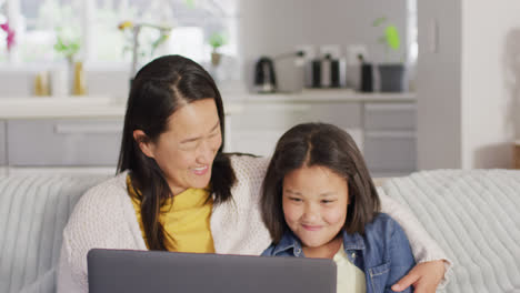 Video-of-happy-asian-mother-and-daughter-sitting-on-sofa-with-laptop