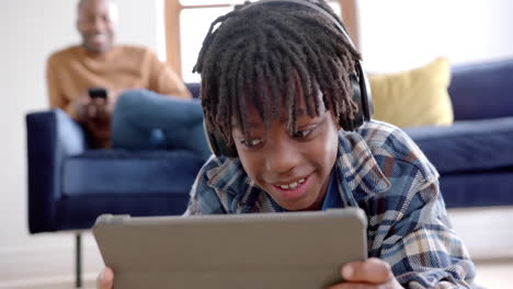 Happy-african-american-boy-with-headphones-using-tablet-lying-on-floor-at-home,-slow-motion