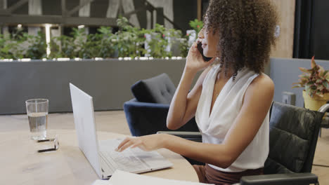 Happy-biracial-businesswoman-in-wheelchair-using-laptop-and-talking-on-smartphone,-in-slow-motion