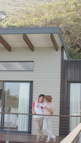 Vertical-video-of-happy-diverse-gay-male-couple-embracing-on-balcony-at-home,-slow-motion