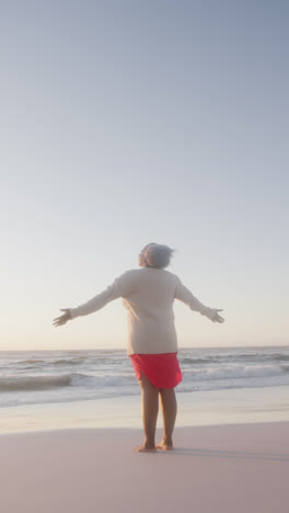 Video-Vertical-De-Una-Feliz-Anciana-Afroamericana-Ampliando-Los-Brazos-En-La-Playa,-En-Cámara-Lenta