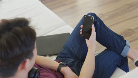 Happy-asian-male-teenager-wearing-headphones-and-using-smartphone-in-living-room