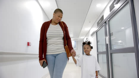 Madre-E-Hija-Afroamericanas-En-Bata-De-Hospital-Cogidas-De-La-Mano-Caminando-En-El-Pasillo,-Cámara-Lenta