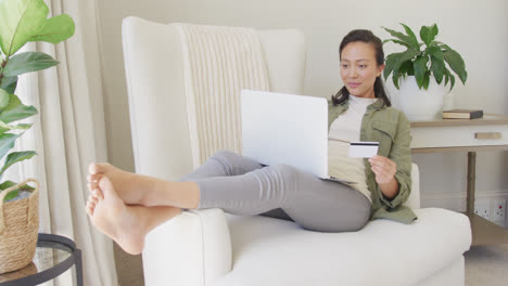 Mujer-Asiática-Feliz-Usando-Una-Computadora-Portátil-Y-Sosteniendo-Una-Tarjeta-De-Crédito-En-El-Dormitorio,-En-Cámara-Lenta