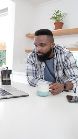 Video-Vertical-De-Un-Hombre-Afroamericano-Tomando-Café-Y-Usando-Una-Computadora-Portátil-En-La-Cocina,-Cámara-Lenta