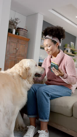 Video-Vertical-De-Una-Mujer-Birracial-Usando-Un-Teléfono-Inteligente-Y-Acariciando-A-Un-Perro-En-Casa,-Cámara-Lenta