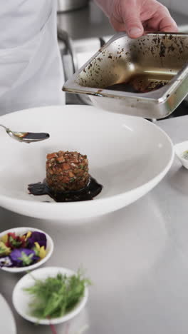 Caucasian-male-chef-pouring-sauce-on-meal-on-white-plate-in-kitchen,-slow-motion,-vertical