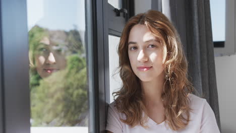 Happy-caucasian-woman-looking-by-window-and-smiling-in-sunny-house,-copy-space