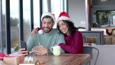 Feliz-Pareja-Birracial-Usando-Sombreros-De-Santa-Claus-Usando-Un-Teléfono-Inteligente-Para-Videollamadas-En-Casa,-Cámara-Lenta