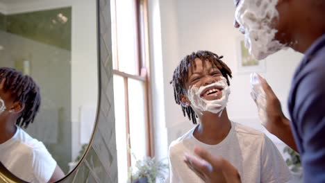 Happy-african-american-father-and-son-applying-shaving-foam-on-face-in-bathroom-at-home,-slow-motion