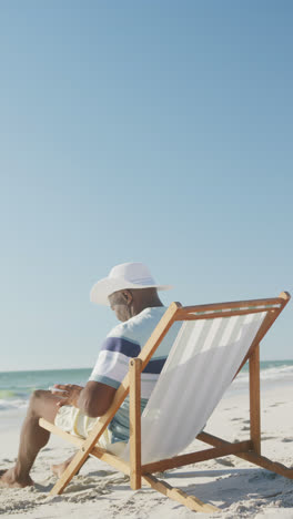 Vertikales-Video-Eines-älteren-Afroamerikanischen-Mannes,-Der-Einen-Laptop-Auf-Einem-Liegestuhl-Am-Strand-Benutzt,-In-Zeitlupe