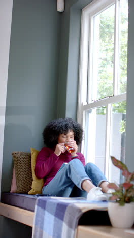 Vertical-video-of-biracial-woman-drinking-tea-and-reading-book-at-home,-slow-motion