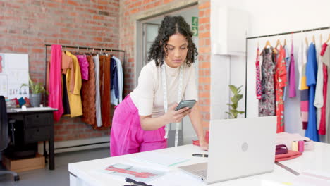 Diseñadora-De-Moda-Birracial-Usando-Una-Computadora-Portátil-Y-Hablando-Por-Teléfono-Inteligente-En-El-Estudio,-Cámara-Lenta