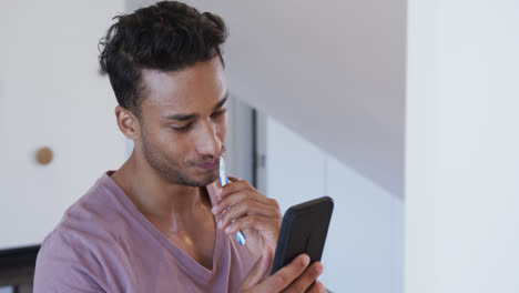 Biracial-man-brushing-teeth-in-bathroom-in-the-morning-and-using-smartphone,-slow-motion