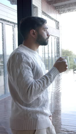 Vertical-video-of-biracial-man-looking-through-window-and-drinking-coffee-at-home,-slow-motion