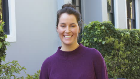 Happy-caucasian-woman-in-purple-sweater-standing-in-front-of-house-in-garden