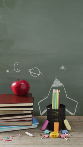 Vertical-video-of-apple,-schoolbooks-and-stationery-on-desk-with-chalk-board-in-background