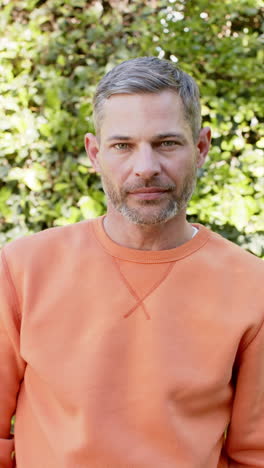 Vertical-video-of-happy-caucasian-man-with-orange-blouse-standing-and-smiling-in-sunny-garden